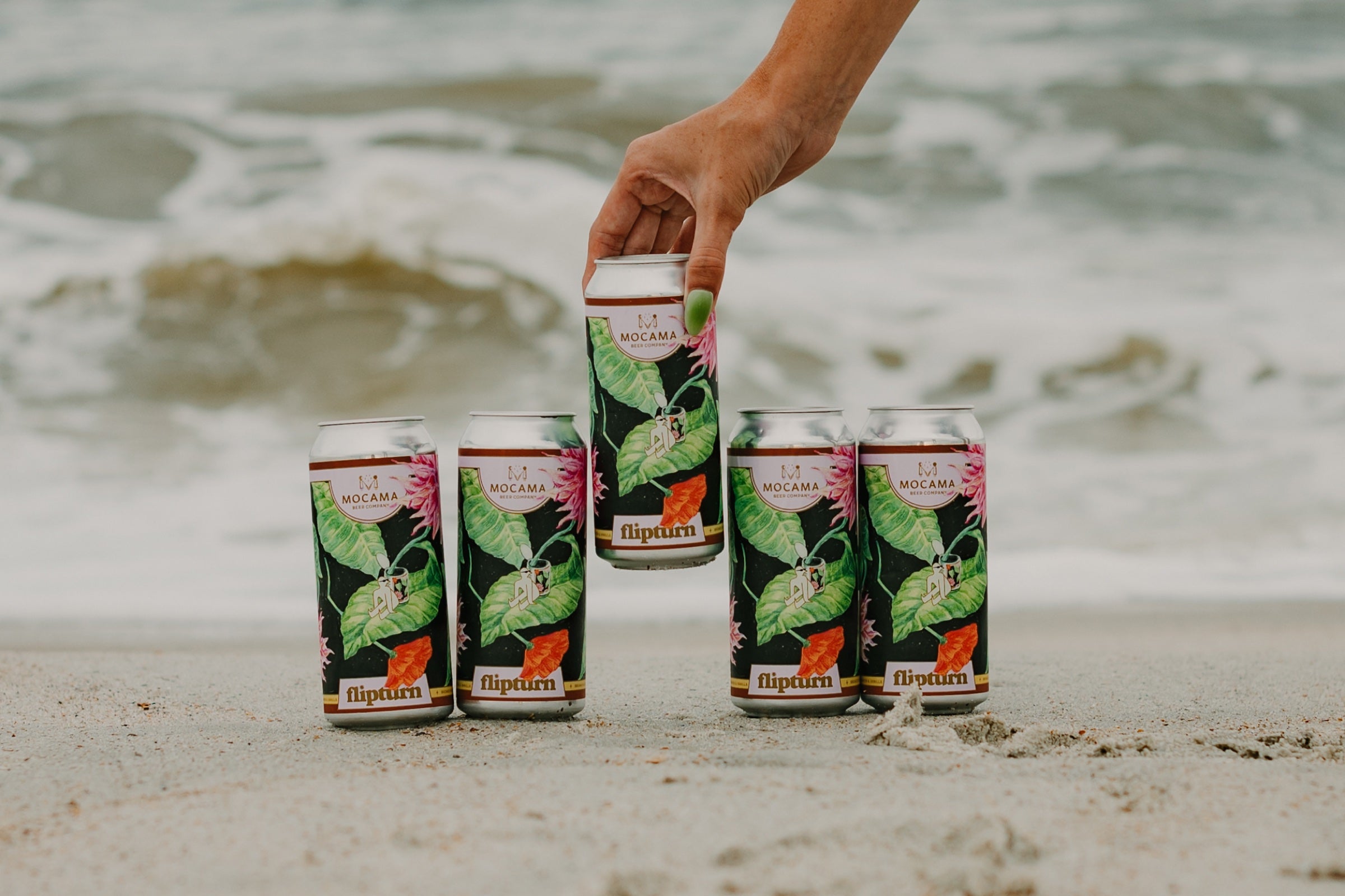 Five cans of Mocama flipturn blonde ale arranged on the beach